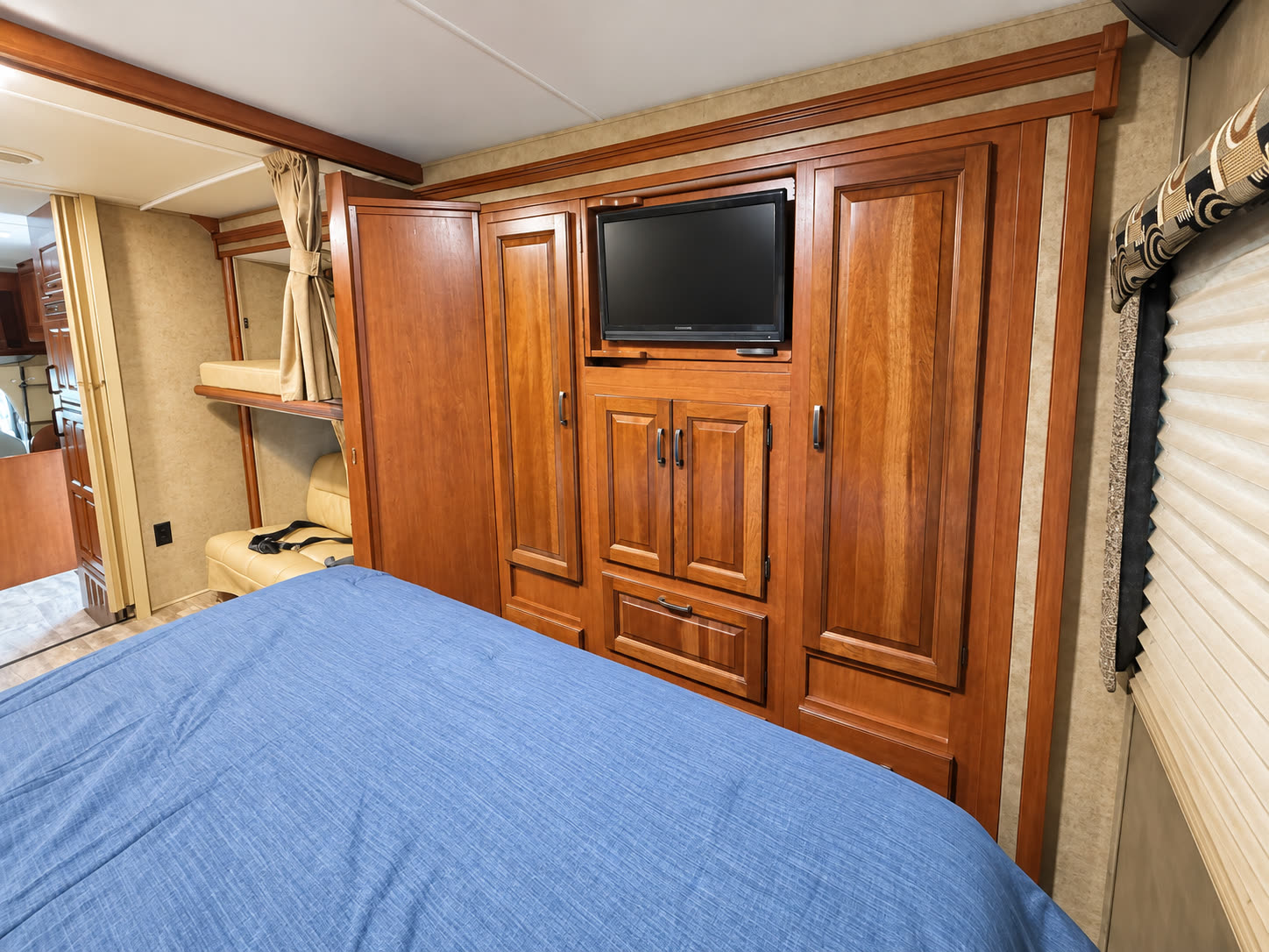 RV bedroom with blue bedspread, large wood wardrobe, mounted TV, and window shade.