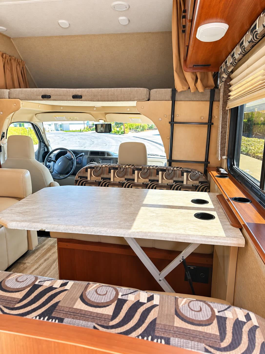 RV dinette with patterned bench seating and table, facing the cab beneath the overhead bunk.