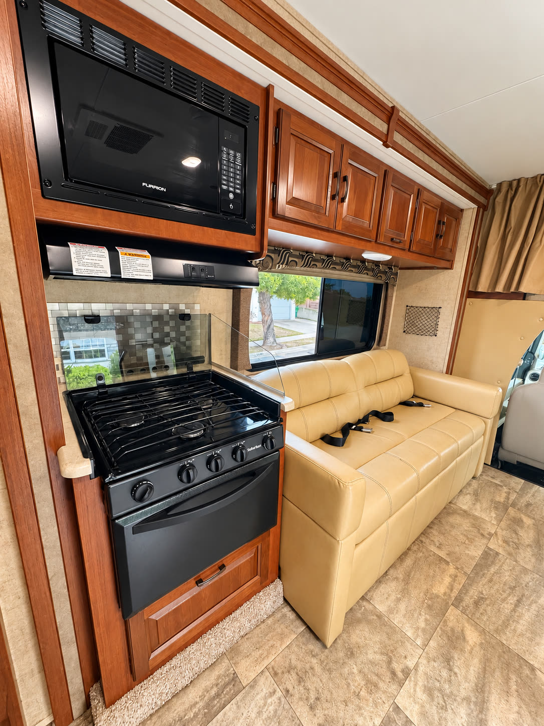 RV galley with black gas range and microwave beside a tan sofa and wood cabinets.