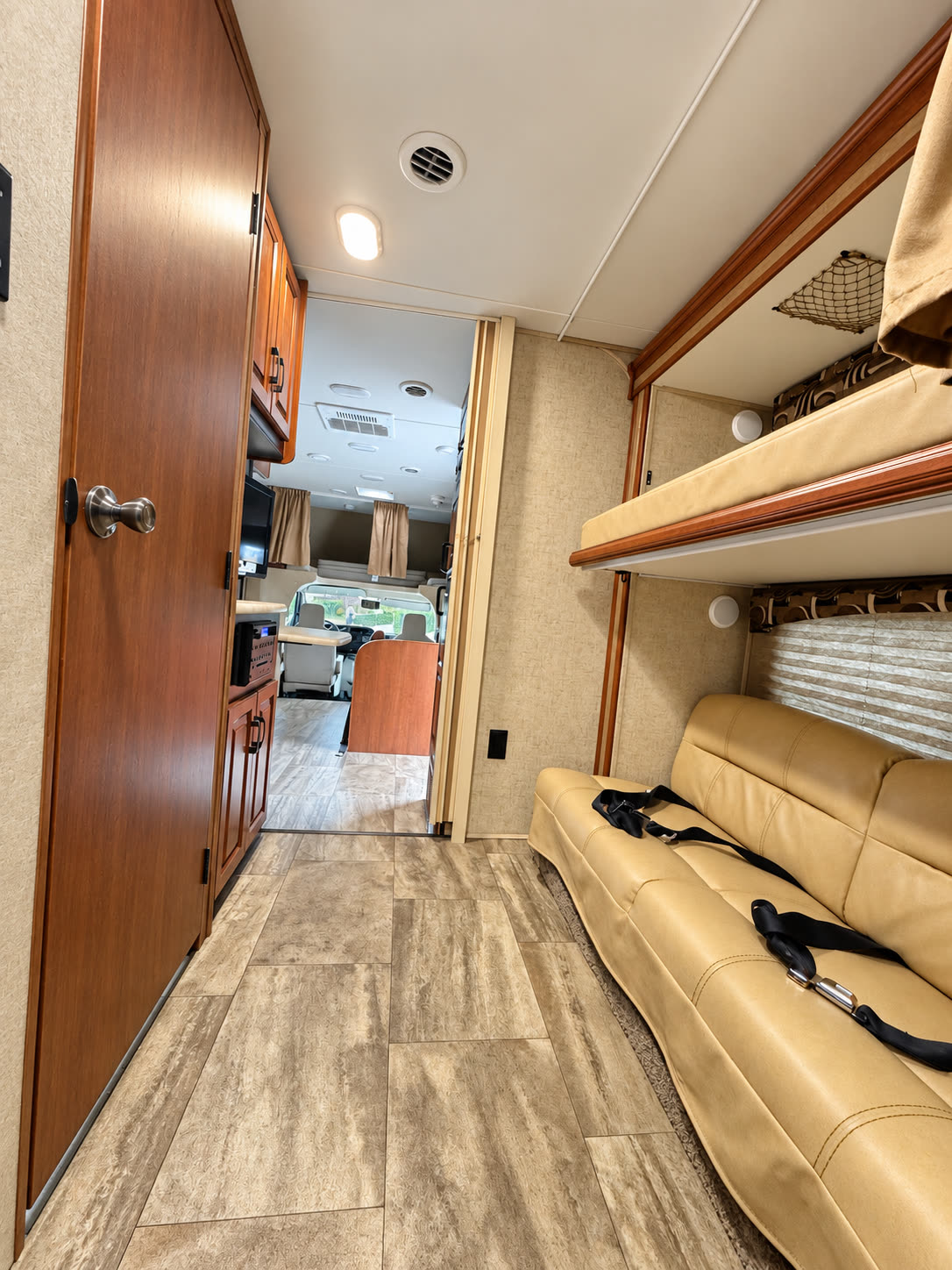 RV interior hallway with tan sofa, overhead bunk, wood cabinetry, and the cab visible ahead.