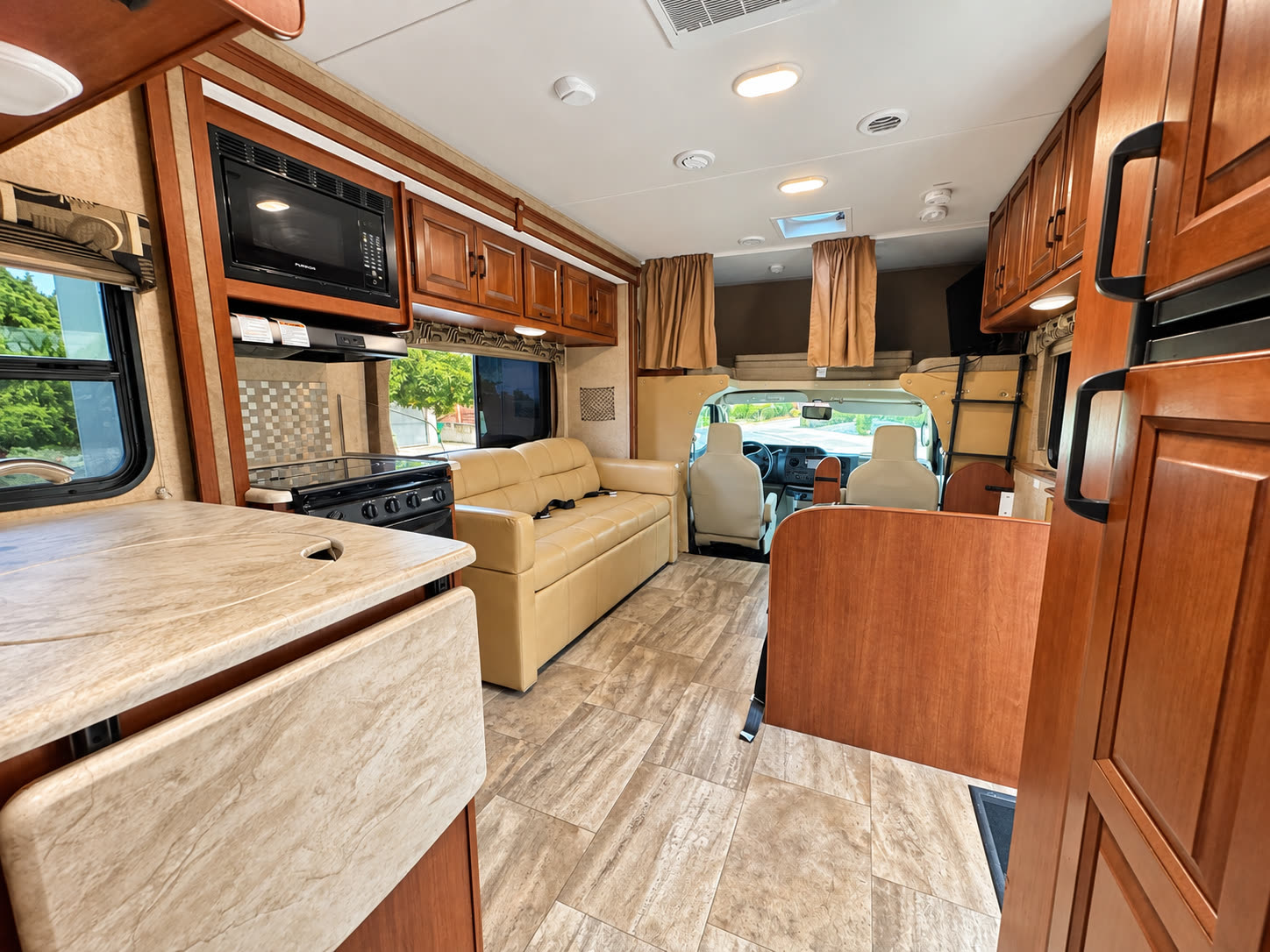 Wide RV interior view showing the kitchen counter, tan sofa, cab, and wood storage cabinets.