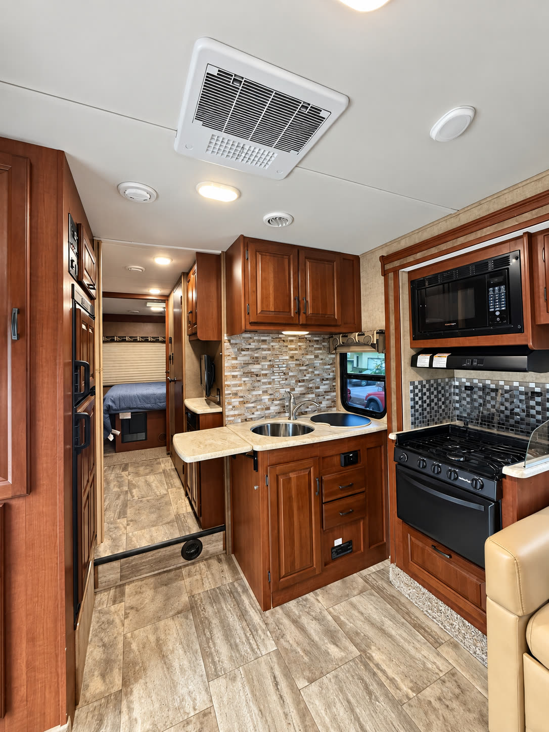 Wide RV interior view of the kitchen, sink, stove, wood cabinetry, and hallway to the bedroom.