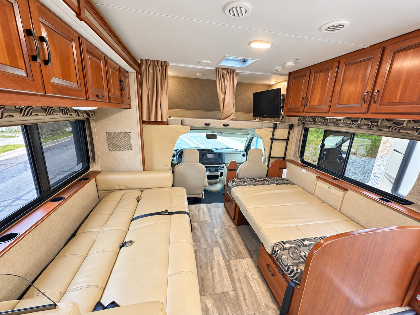 RV living area with tan sofa, convertible dinette, cab-over bed, wood cabinets, and side windows.