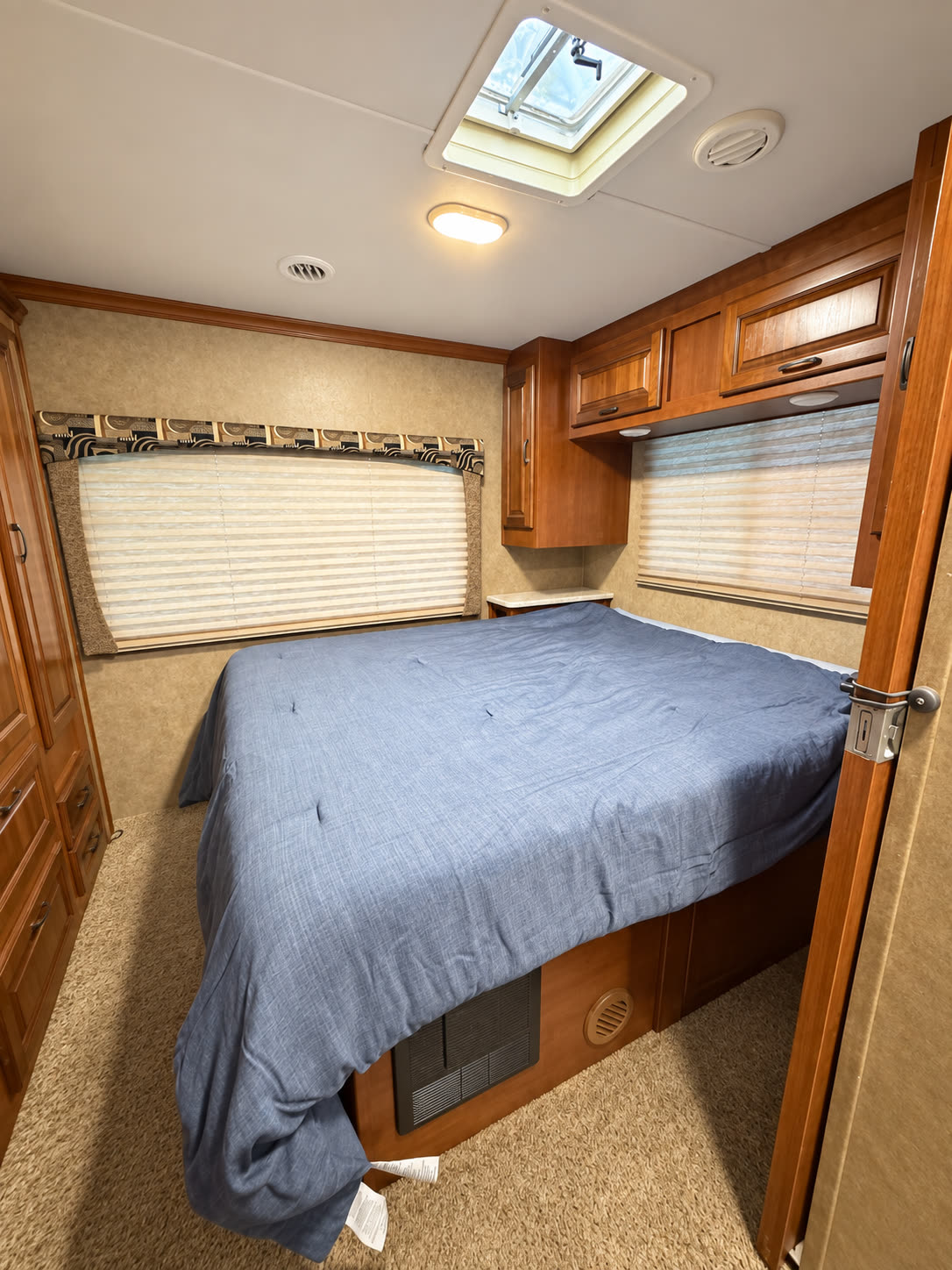 Rear RV bedroom with blue-covered bed, overhead wood cabinets, windows, and skylight.